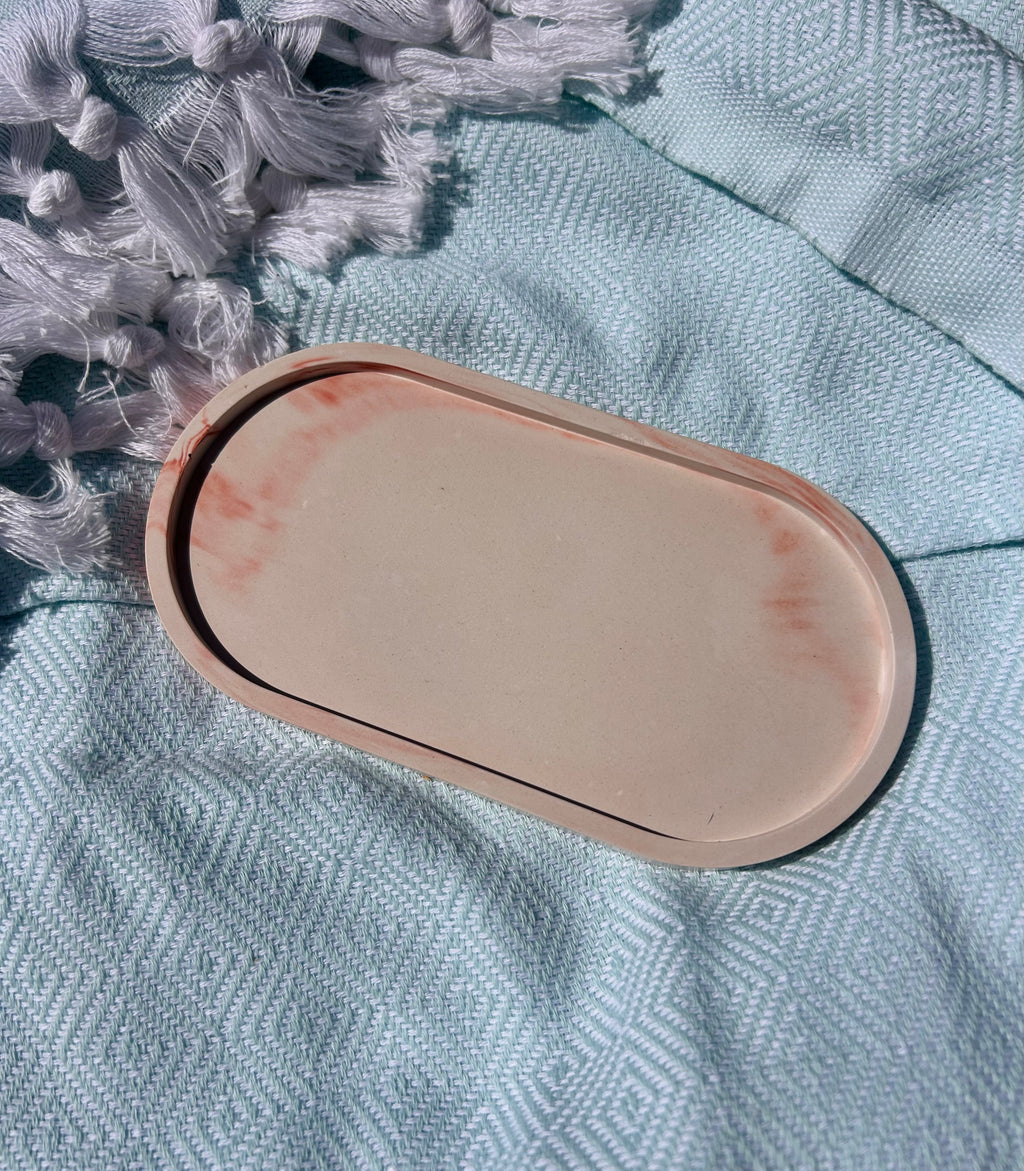 An oval resin tray with a light pink border and a cream center, featuring subtle terracotta-colored swirls near the edges, resting on a textured light blue fabric surface.