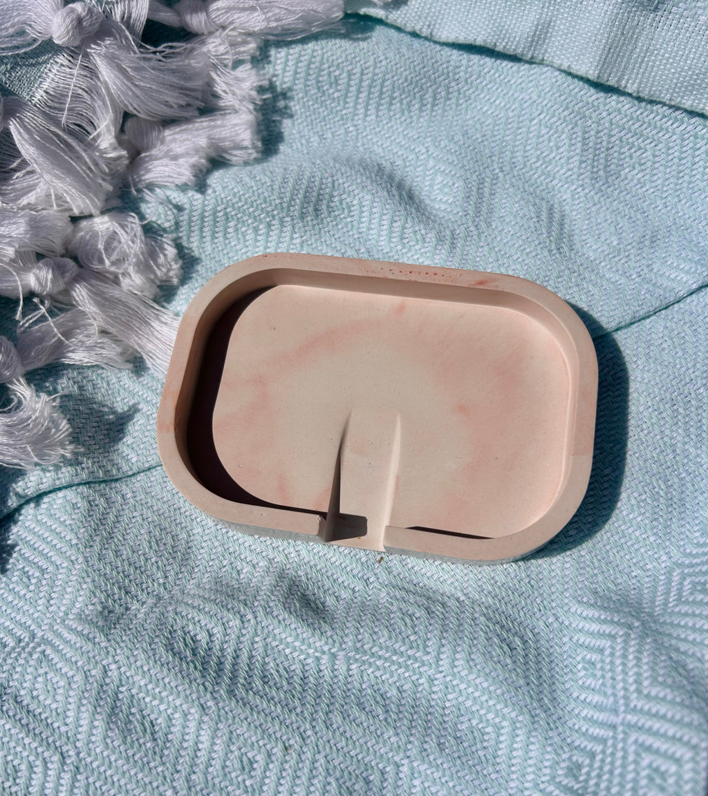 A small rounded rectangular tray or dish in a pale peach-beige color with a central divider, placed on a textured light blue fabric surface with white tassels visible in the upper left.