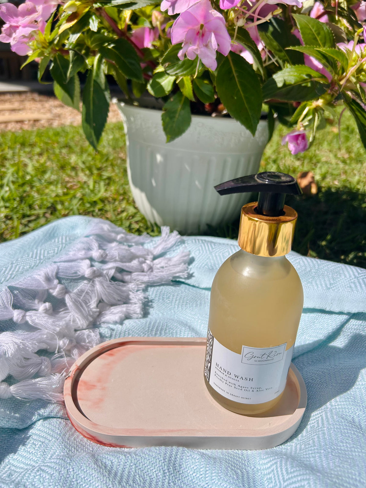 A hand wash bottle with a gold collar and black pump sits on a peach tray on a light blue blanket outdoors, with a potted pink flower in the background.