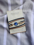 A Muga Muga folk jewellery bracelet with a blue and white patterned central cabochon and gold-toned hardware, wrapped in tan and navy cords, displayed on its cream-colored branded card over a gray and white striped fabric background.