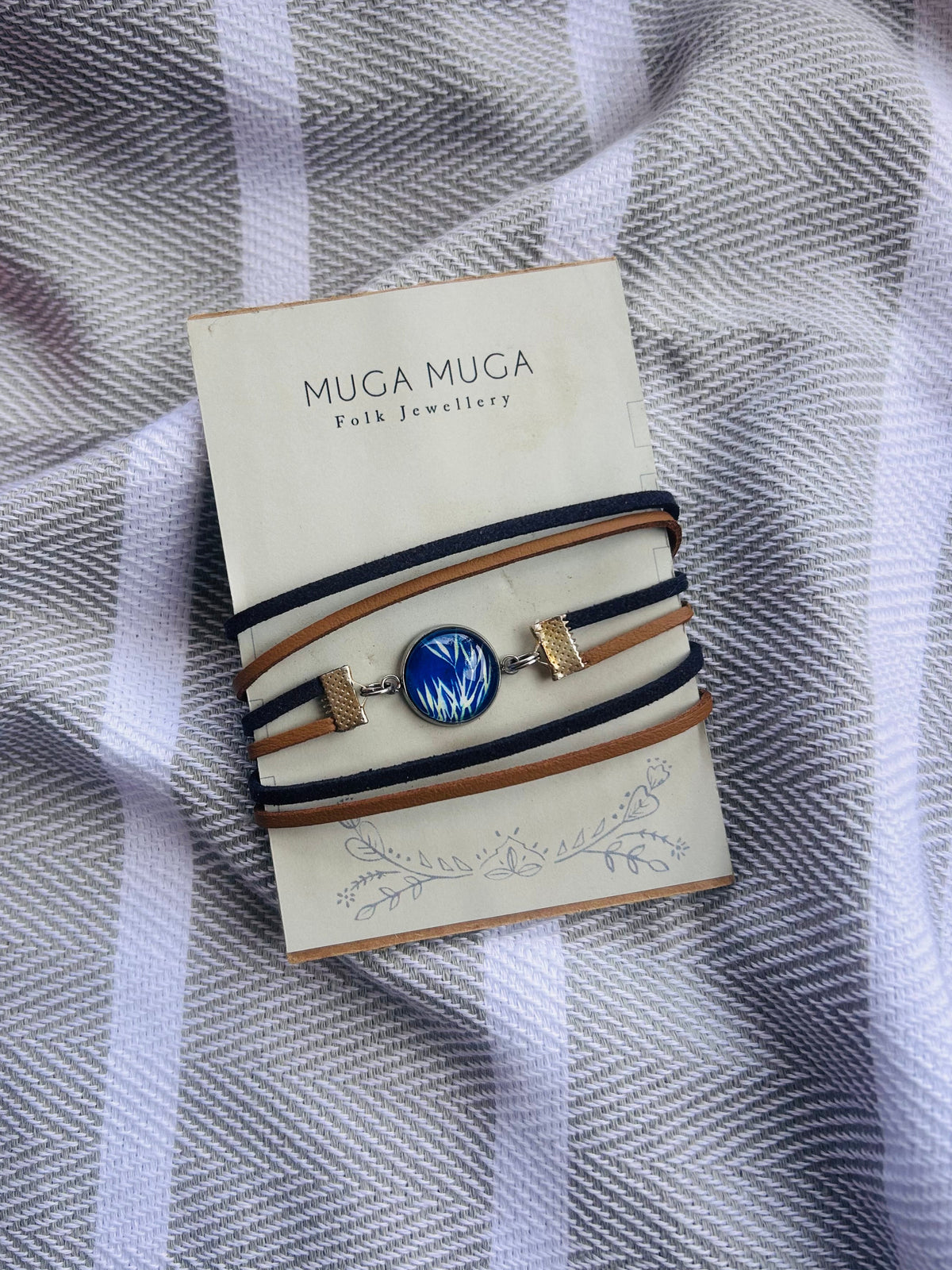A Muga Muga folk jewellery bracelet with a blue and white patterned central cabochon and gold-toned hardware, wrapped in tan and navy cords, displayed on its cream-colored branded card over a gray and white striped fabric background.