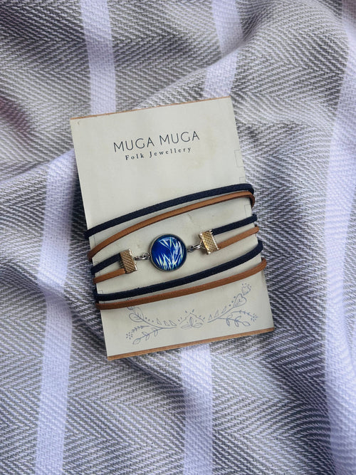 A Muga Muga folk jewellery bracelet with a blue and white patterned central cabochon and gold-toned hardware, wrapped in tan and navy cords, displayed on its cream-colored branded card over a gray and white striped fabric background.