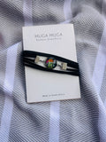 A black multi-strand wrap bracelet with a colorful floral cabochon and silver-tone end pieces, displayed on a white MUGA MUGA card over a gray and white striped fabric background.