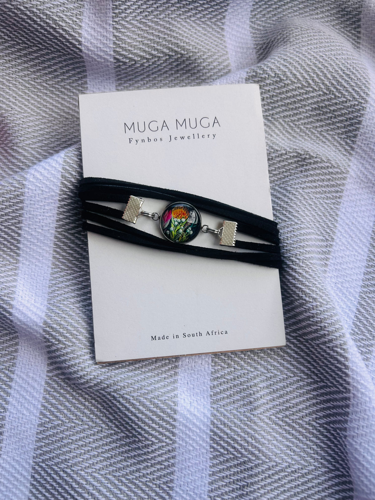 A black multi-strand wrap bracelet with a colorful floral cabochon and silver-tone end pieces, displayed on a white MUGA MUGA card over a gray and white striped fabric background.