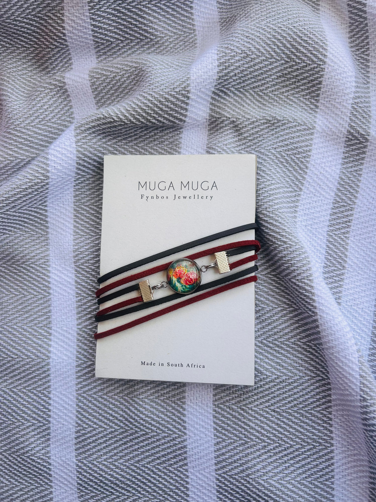 A Muga Muga Jewellery bracelet with a colorful, iridescent round cabochon and gold-toned metal accents, wrapped in black, red, and white cords. The card is displayed on a gray and white striped textured fabric background.