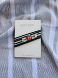 A black multi-strand cord bracelet with a colorful mosaic-style oval centerpiece and small gold-toned rectangular accents, displayed on a cream card labeled MUGA MUGA Jewellery, placed on a gray-and-white striped fabric background.