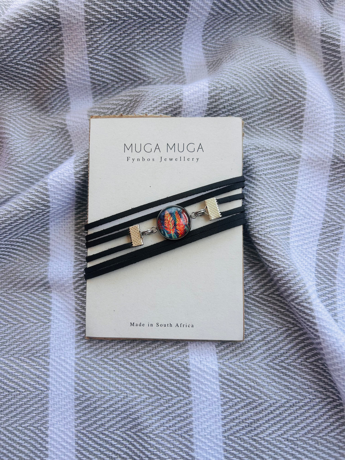 A black multi-strand cord bracelet with a colorful mosaic-style oval centerpiece and small gold-toned rectangular accents, displayed on a cream card labeled MUGA MUGA Jewellery, placed on a gray-and-white striped fabric background.