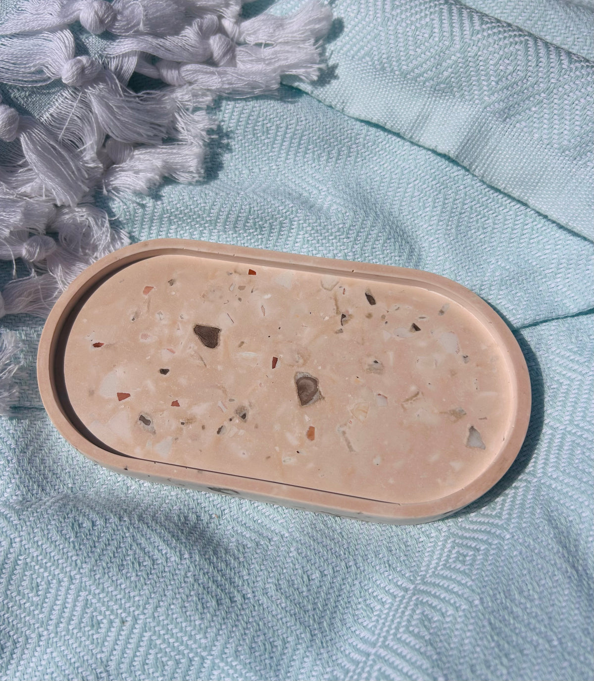 An oval-shaped beige tray with a terrazzo-style speckled pattern, resting on a textured light blue fabric surface next to white tassels.