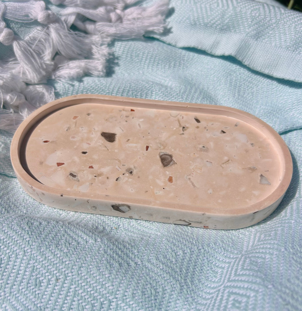 An oval concrete tray with a terrazzo-like speckled pattern in beige, white, and brown, resting on a textured light blue blanket.