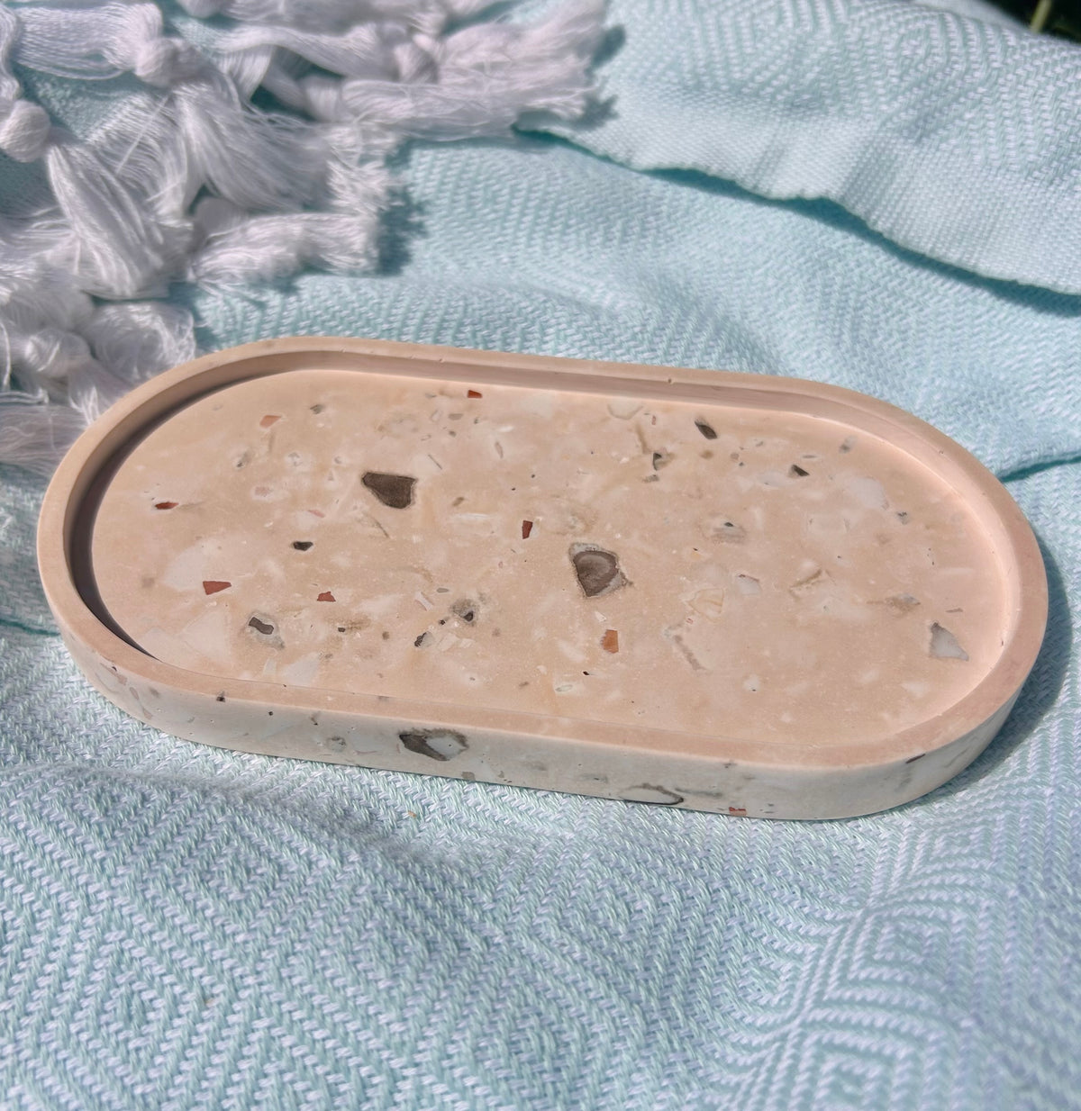 An oval concrete tray with a terrazzo-like speckled pattern in beige, white, and brown, resting on a textured light blue blanket.