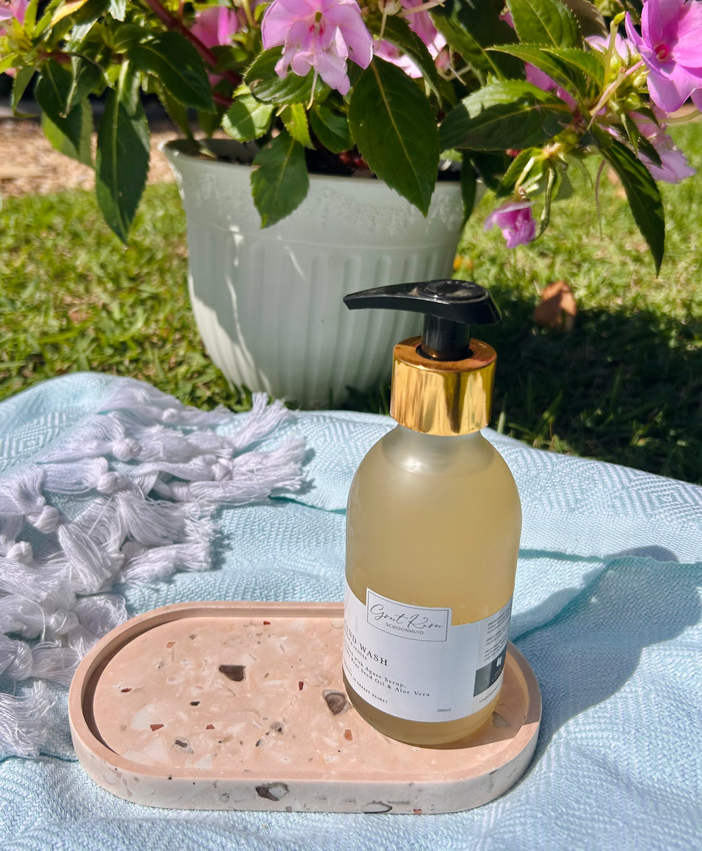 A frosted glass pump bottle with a black pump and gold collar sits on a peach stone tray outdoors on a light blue blanket, with grass, flowers, and a white planter in the background.