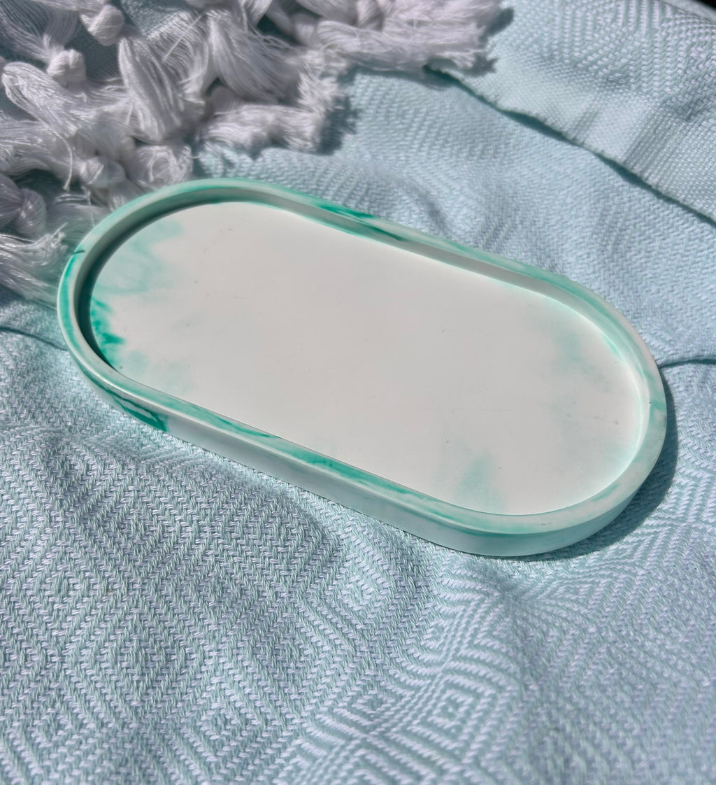 An oval resin tray with a white center and mint green marbled edges, resting on a textured light blue fabric surface with white tassels in the background.