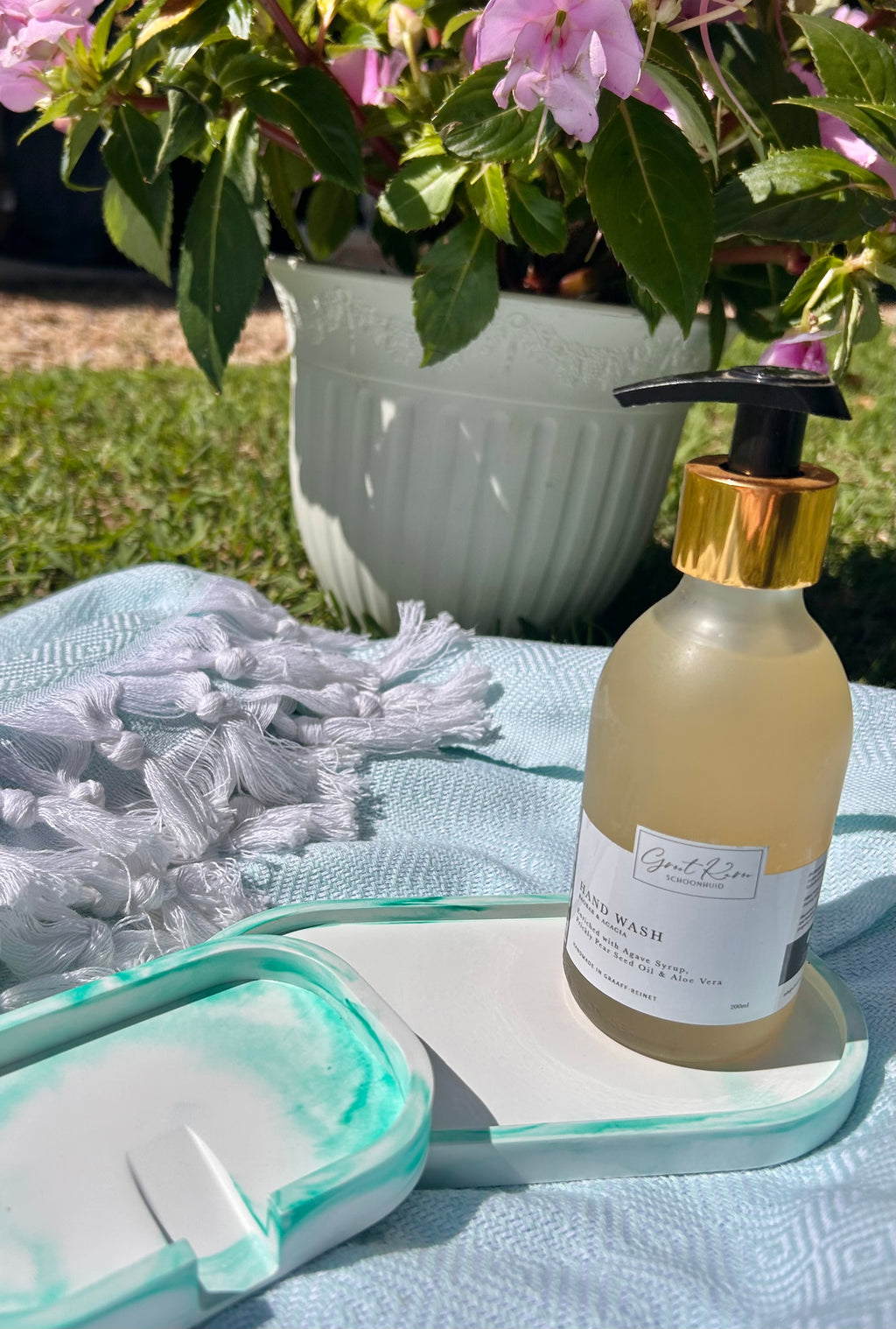 A bottle of hand wash with a gold collar and black pump sits on a white tray on a light blue blanket outdoors, with a potted plant of pink flowers in the background.