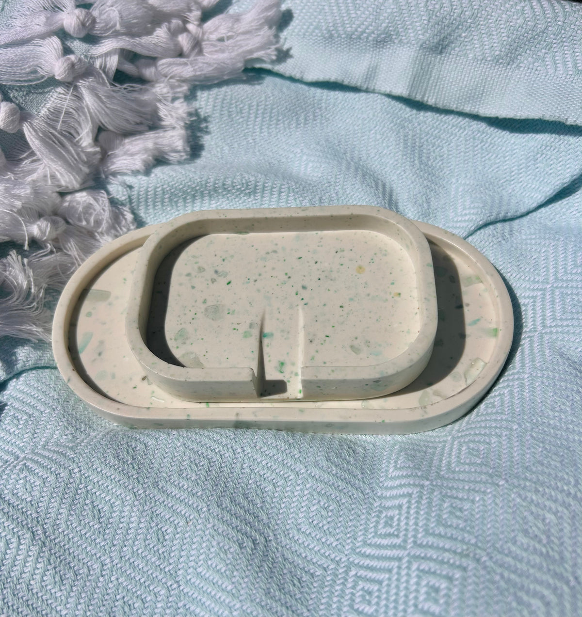 A speckled cream-colored soap dish with a matching inner tray, placed on a textured light blue blanket with white tassels in the corner.