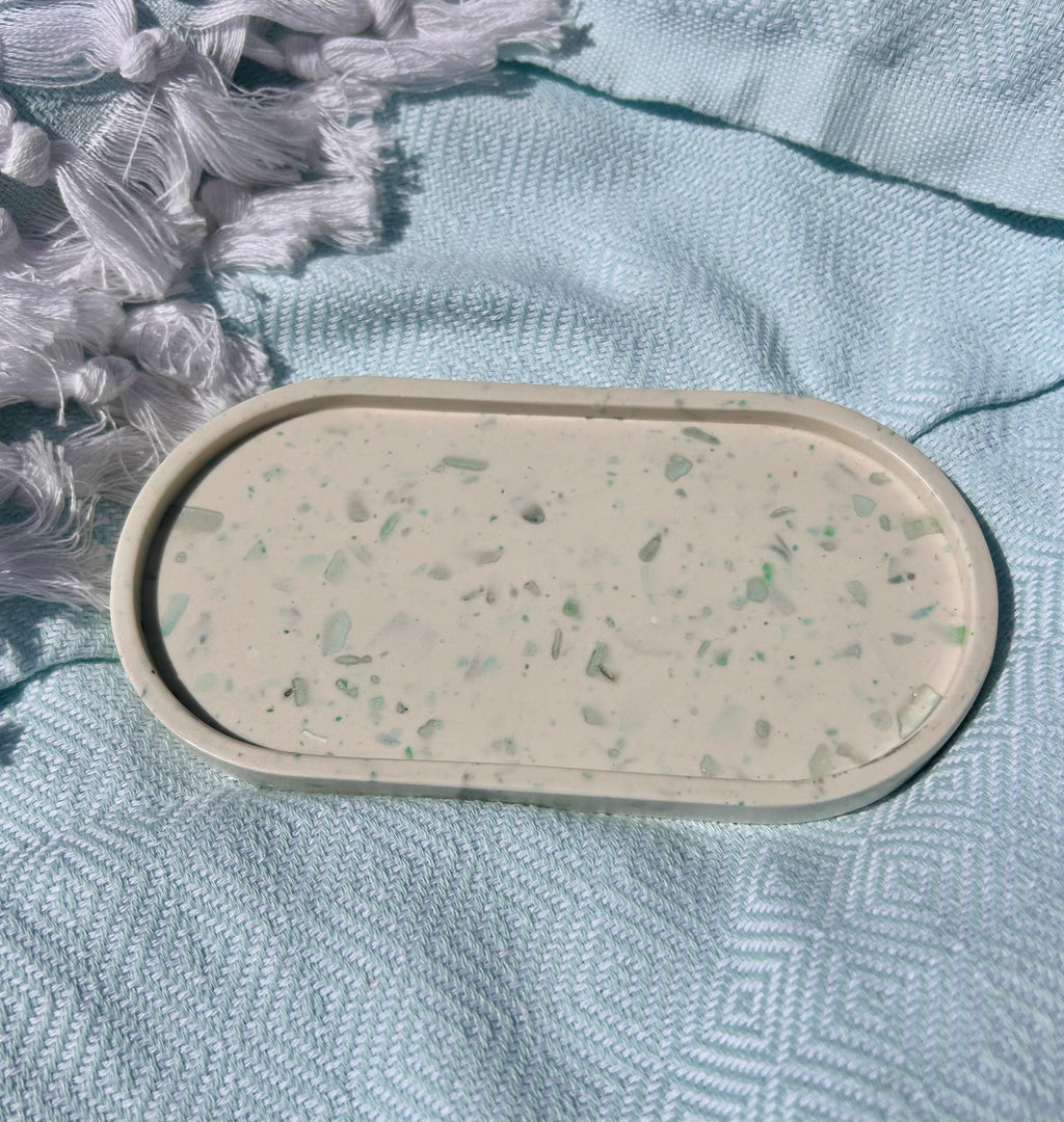An oval terrazzo-style soap dish or tray in a warm cream color with small green and gray flecks, resting on a textured light blue blanket.
