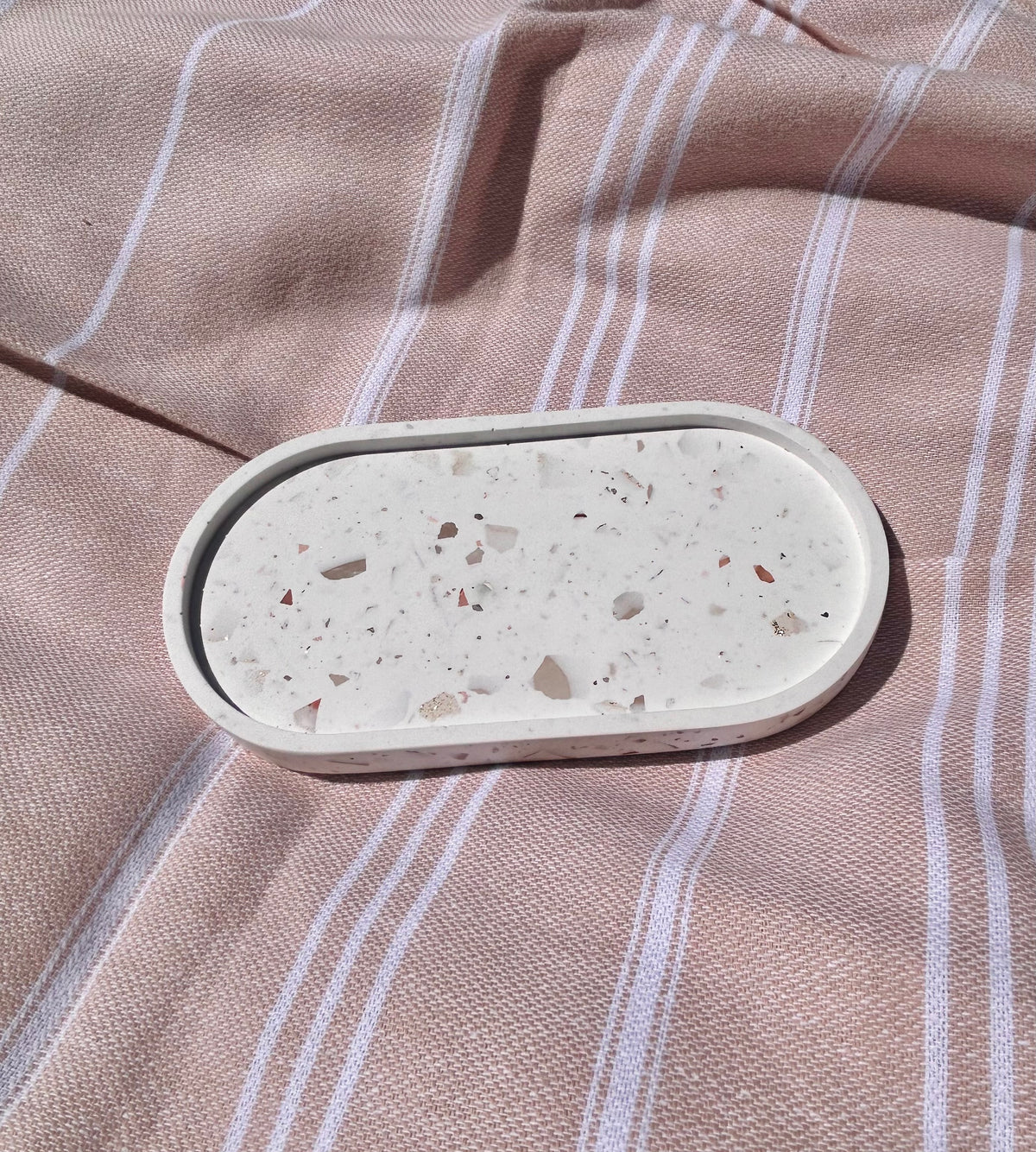 An oval terrazzo tray with a speckled cream and brown pattern, resting on a dusty pink and white striped fabric background.