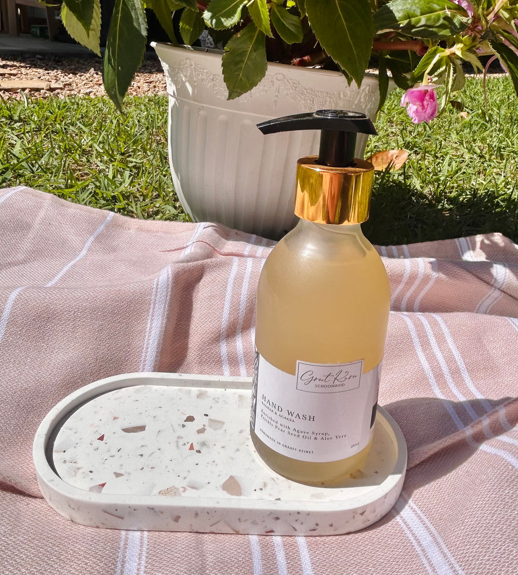 A bottle of hand wash with a black pump and gold collar sits on a white tray on a pink striped blanket outdoors, with grass and a potted plant in the background.