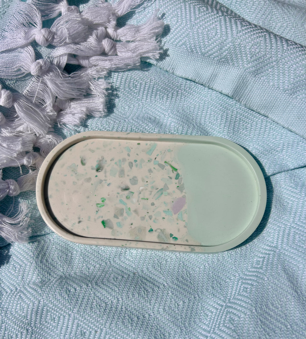 An oval handmade soap bar with a half-and-half design, resting on a textured light blue fabric next to white tassels.