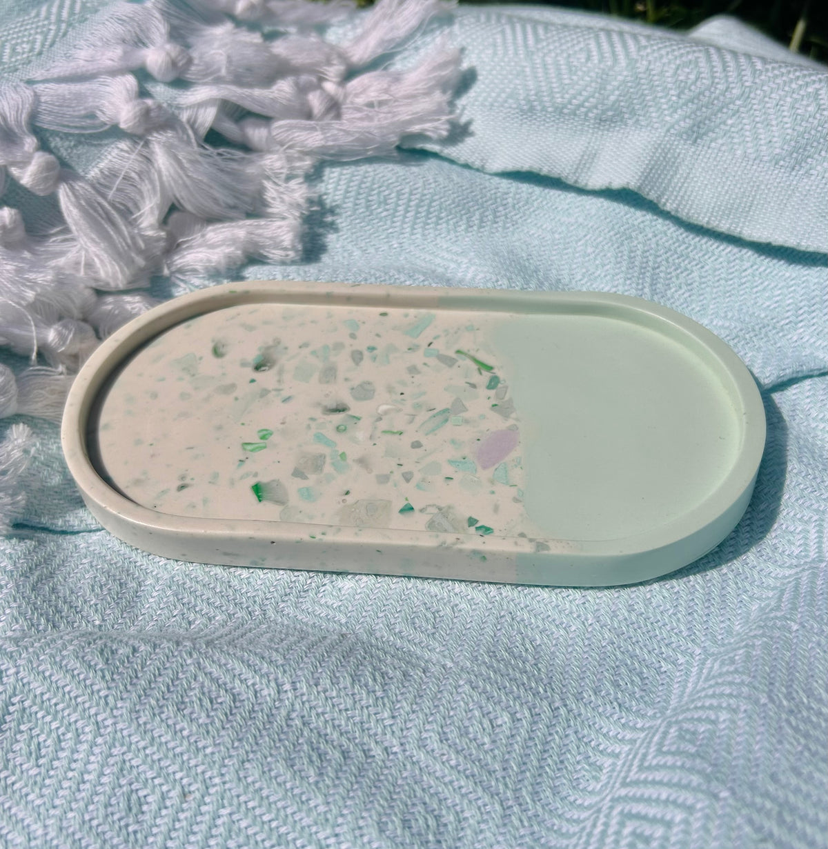 An oval resin tray with a half-painted sage green finish and a half terrazzo pattern, resting on a textured light blue blanket.