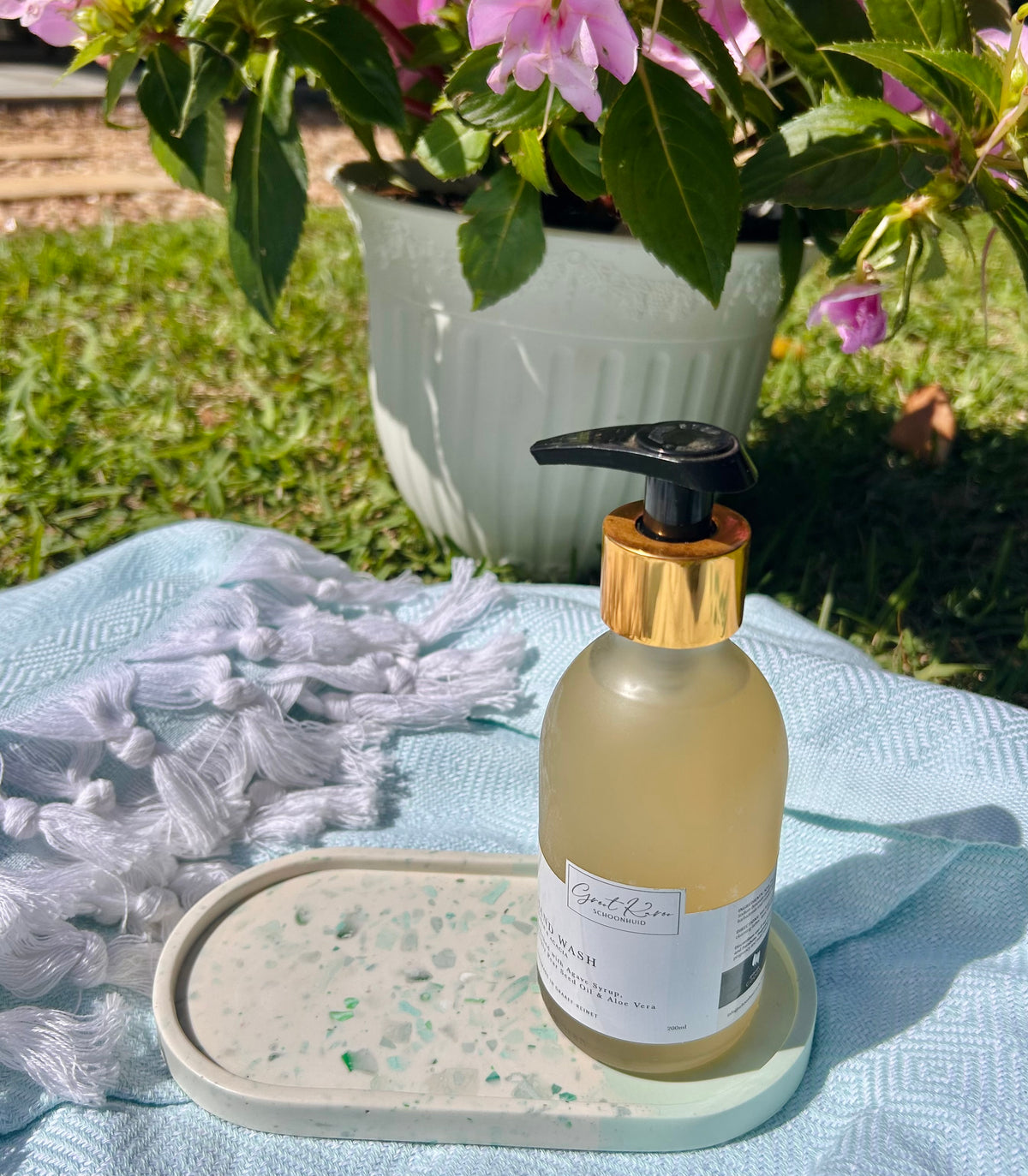 A frosted glass pump bottle with a gold collar and black pump, filled with pale yellow liquid, sits on a light blue textured blanket outdoors. The bottle is displayed on a speckled tray next to white tassels, with a potted plant of pink flowers in the background.
