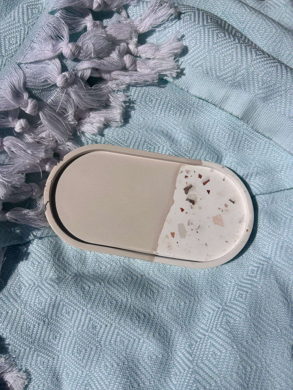 An oval-shaped ceramic dish or coaster with a split design, featuring a solid beige half and a terrazzo-style white half with small pink and brown chips, resting on a textured light blue fabric with gray tassels.