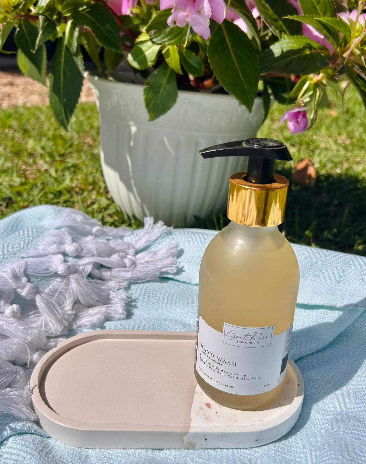 A bottle of hand wash with a black pump and gold collar sits on a small tray on a light blue blanket outdoors, with a potted plant of pink flowers in the background.