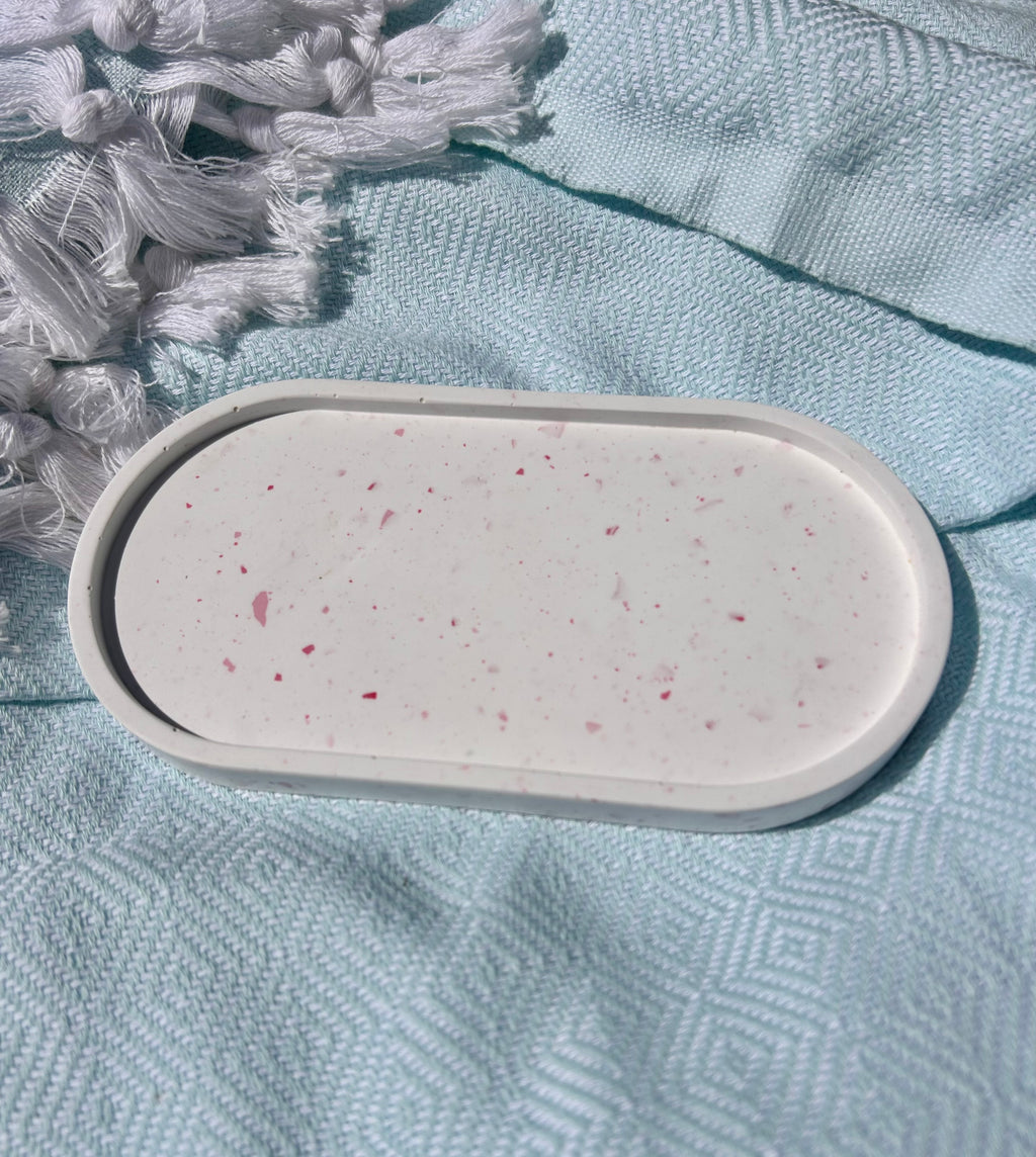 An oval white tray with a raised rim, speckled with small pink and red flecks, resting on a textured light blue fabric surface next to white tassels.