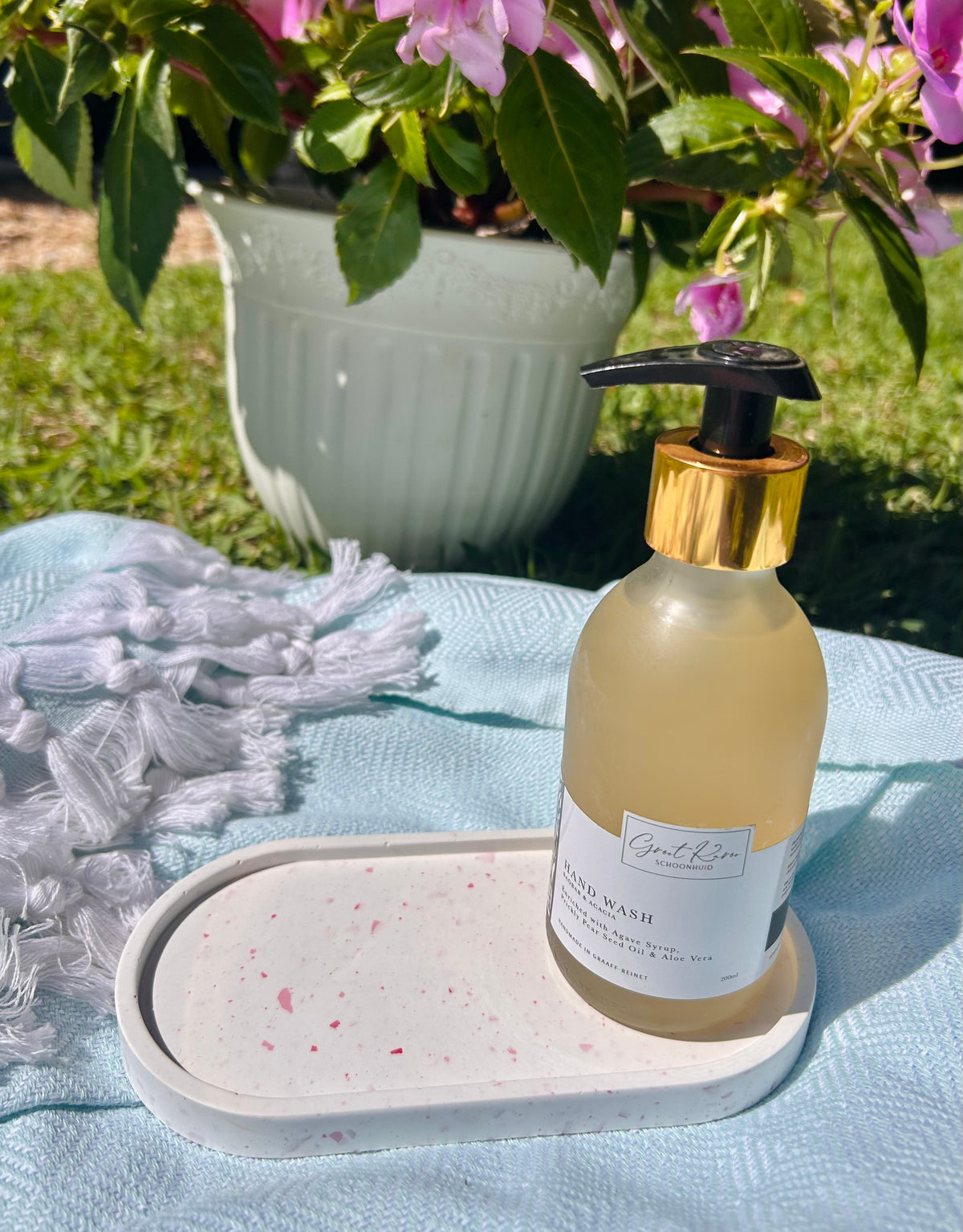 A bottle of hand wash with a black pump and gold collar, sitting on a white speckled tray on a light blue blanket outdoors with flowers and grass in the background.