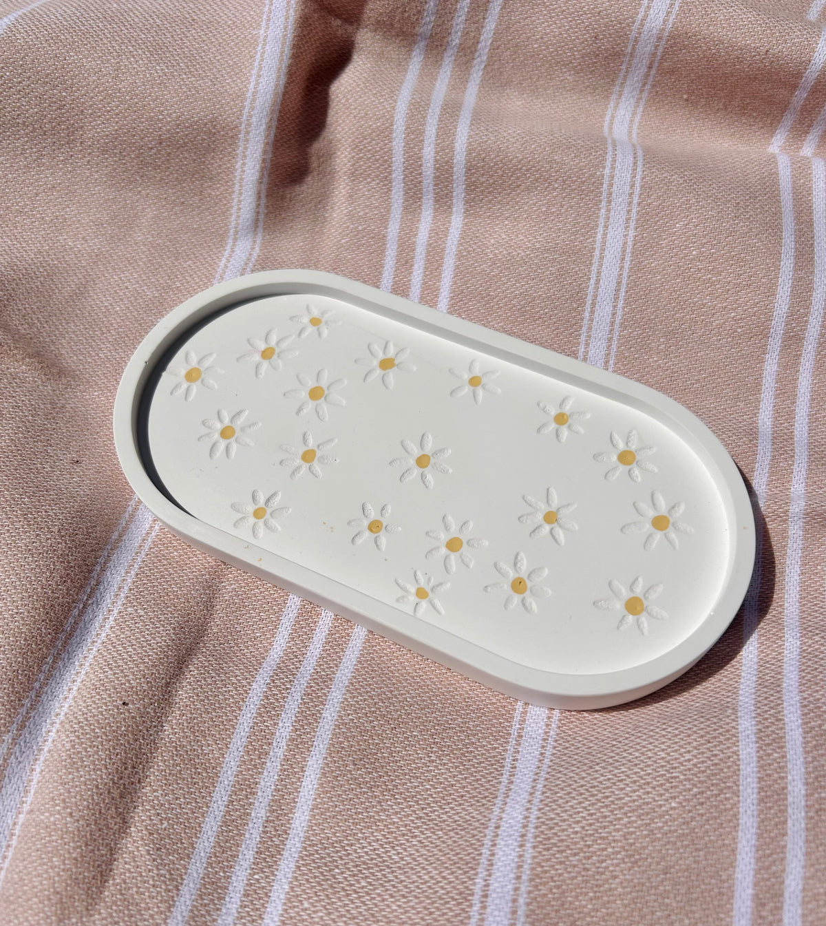 An oval cream-colored silicone tray with a daisy pattern, placed on a pink and white striped fabric background.